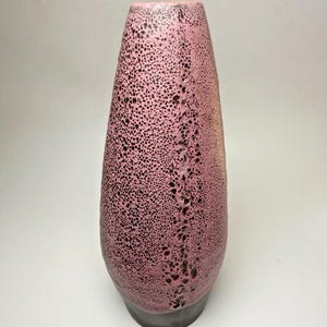 May include: A tall, pink ceramic vase with a speckled pattern and a dark grey base. The vase has a textured surface with small, dark spots and a vertical line of larger, darker holes. The vase is approximately 30 cm tall.