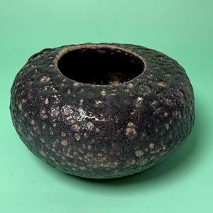 May include: A dark, textured ceramic bowl with a wide opening. The bowl is a deep, mottled black with speckled, lighter-colored accents. The rim has a rough, uneven texture. The bowl sits on a solid green surface.