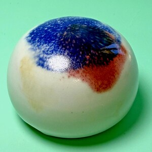 May include: A smooth, dome-shaped ceramic paperweight with a glossy finish. The base color is off-white, featuring a prominent abstract splash of deep blue and reddish-brown pigment on the top surface, creating a distinct, mottled color effect.