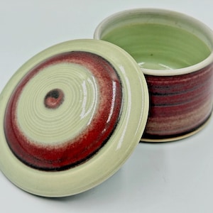 May include: A cylindrical ceramic jar with a matching lid, featuring a glossy glaze in shades of pale mint green and deep burgundy. The lid displays concentric circular patterns, while the jar body has horizontal bands of color.