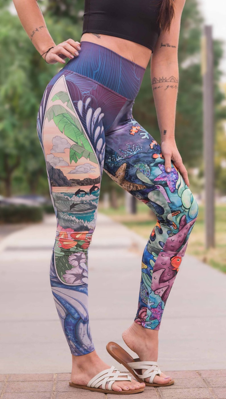 May include: A pair of colorful leggings with a tropical and underwater print. The leggings feature a blue wave, palm trees, fish, and coral. The leggings are worn by a person with tattoos on their arms.