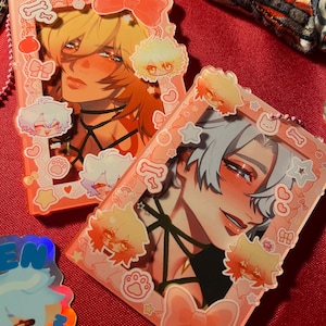 以下が含まれることがあります： 2つのピンクのアクリルチャーム。アニメスタイルの男性キャラクターのイラストが描かれています。各チャームには、リボン、足跡、骨の形をしたピンク色のフレームが付いています。1つは金髪のキャラクター、もう1つは銀髪のキャラクターです。