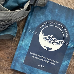 May include: A blue journal with the text "Adirondack High Peaks" and a white mountain range illustration. The text below the illustration reads "A trail journal for conquering the 46 Adirondack High Peaks".