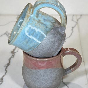 Puede incluir: Dos tazas de cerámica hechas a mano apiladas una encima de la otra. La taza superior es azul claro y gris, mientras que la inferior es gris con una banda rosa. Ambas tazas tienen asas grandes y curvas.