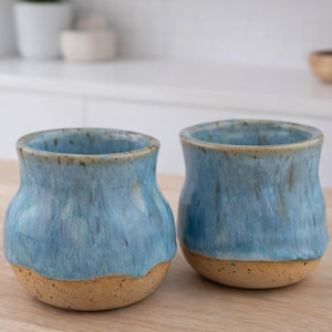 May include: Two handmade ceramic cups. The cups have a light blue glaze over a tan base. The blue glaze has a textured appearance. The cups have a rounded shape and a slightly flared rim. The cups are suitable for beverages.