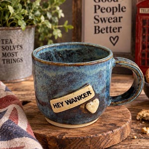Puede incluir: Una taza de cerámica moteada en azul y marrón con asa, que presenta una placa rectangular de color crema con el texto negro "HEY WANKER" y un pequeño corazón esculpido de color crema adherido a la superficie frontal de la taza.