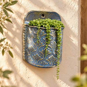 May include: A wall-mounted ceramic planter featuring a deep blue glaze with gold-toned highlights and a repeating embossed floral pattern. The vessel contains a trailing string of pearls succulent with small, round, vibrant green leaves.
