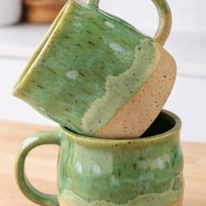 May include: Two handmade ceramic mugs stacked on top of each other. The mugs are glazed in shades of green with a speckled, textured finish. The bottom portion of each mug is a natural, unglazed tan color. Each mug has a rounded handle.