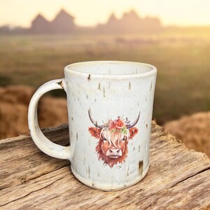 May include: A ceramic mug with a white speckled finish. The mug features a watercolor illustration of a Highland cow with a floral crown. The mug has a curved handle and is set on a wooden surface, with a blurred outdoor background.