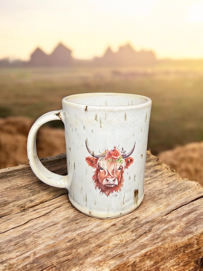 May include: A ceramic mug with a white speckled finish. The mug features a watercolor illustration of a Highland cow with a floral crown. The mug has a curved handle and is set on a wooden surface, with a blurred outdoor background.