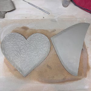 May include: A heart-shaped clay piece with a leaf pattern and a triangular clay piece. The heart is embossed with a detailed leaf design. The clay pieces are on a wooden surface, suggesting a pottery or crafting project.