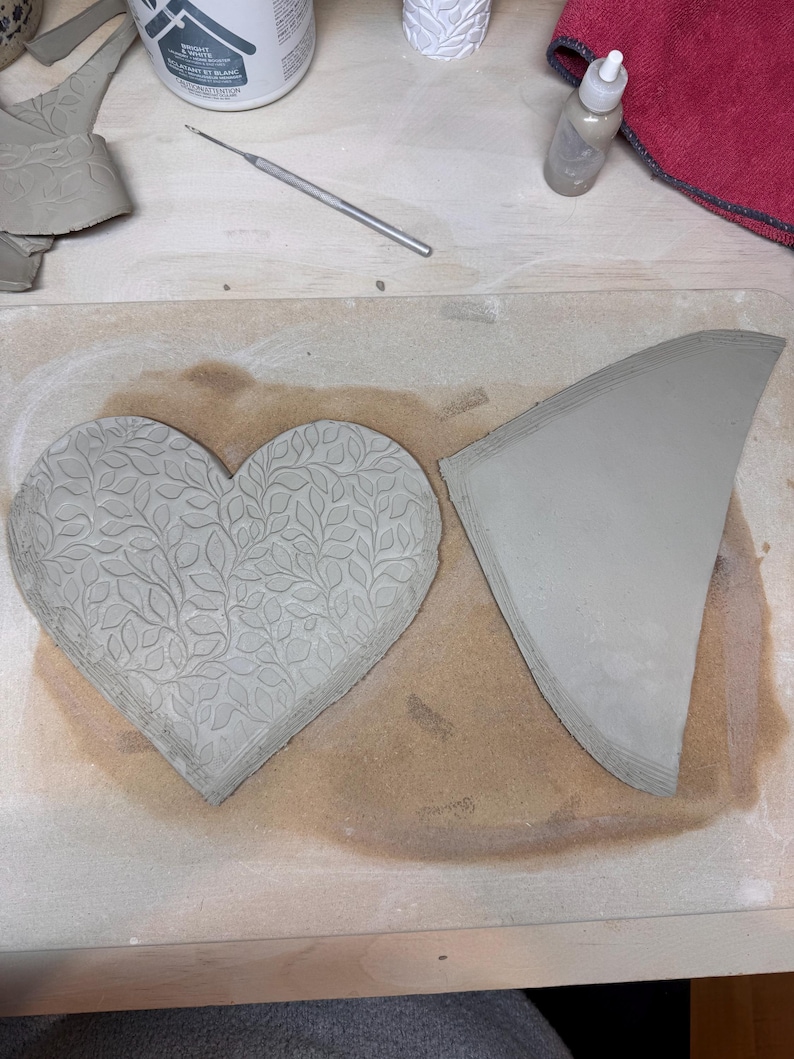 May include: A heart-shaped clay piece with a leaf pattern and a triangular clay piece. The heart is embossed with a detailed leaf design. The clay pieces are on a wooden surface, suggesting a pottery or crafting project.