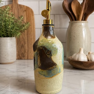 Puede incluir: Una botella dispensadora de aceite de cer&aacute;mica con una base crema moteada y parches abstractos de esmalte marr&oacute;n y azul. La botella cuenta con un vertedor de metal dorado y el texto Walnut - special edition impreso en la parte inferior.