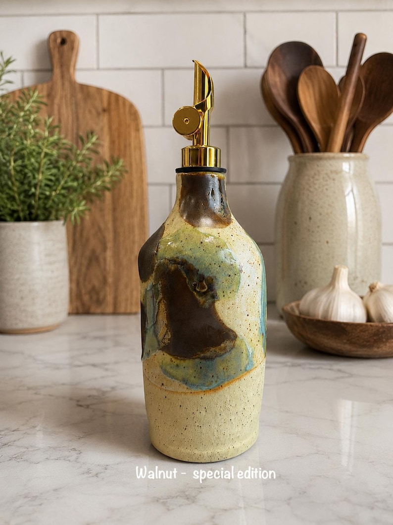 Puede incluir: Una botella dispensadora de aceite de cer&aacute;mica con una base crema moteada y parches abstractos de esmalte marr&oacute;n y azul. La botella cuenta con un vertedor de metal dorado y el texto Walnut - special edition impreso en la parte inferior.
