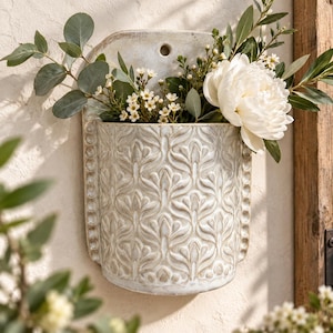 May include: A wall-mounted ceramic pocket planter featuring a cream-coloured finish with a raised, repeating floral relief pattern and a beaded border. The planter holds a bouquet of white peonies, small white flowers, and green eucalyptus leaves.