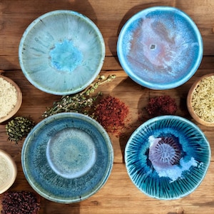 May include: Four ceramic plates in shades of blue and teal with a glossy, textured finish are displayed on a wooden surface. The plates have varied designs, including concentric circles and mottled patterns. Small wooden bowls and spices are also visible.