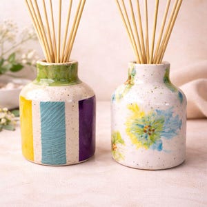 May include: Two ceramic reed diffuser bottles with wooden reeds. One bottle has vertical stripes of yellow, blue, and purple. The other has blue and yellow floral designs. Both have green rims.