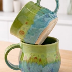 May include: Two handmade ceramic mugs stacked on top of each other. The mugs have a rounded shape with a large handle. The mugs are glazed in shades of green, blue, and brown. The mugs are sitting on a wooden surface.
