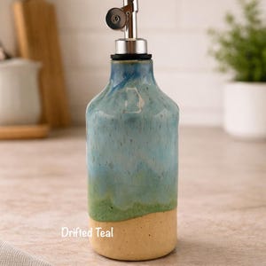 Puede incluir: Una botella dispensadora de aceite de cer&aacute;mica con un esmalte degradado que pasa de una base beige claro a una parte superior verde y azul turquesa. La botella tiene un vertedor de acero inoxidable con tapa y el texto Drifted Teal.