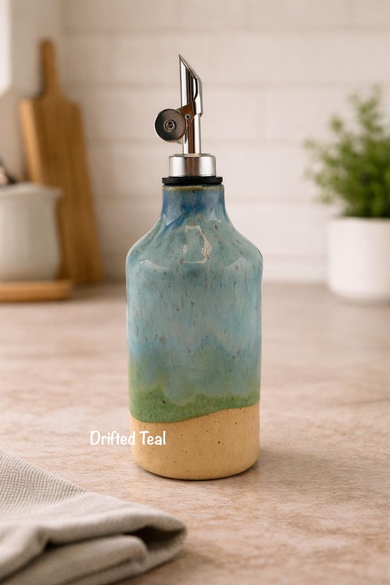 Puede incluir: Una botella dispensadora de aceite de cer&aacute;mica con un esmalte degradado que pasa de una base beige claro a una parte superior verde y azul turquesa. La botella tiene un vertedor de acero inoxidable con tapa y el texto Drifted Teal.