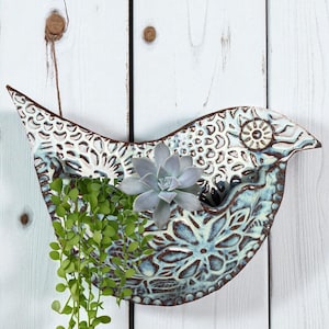 Puede incluir: Un macetero de pared de cerámica con forma de pájaro y diseño floral azul y blanco desgastado. El macetero contiene una suculenta y plantas verdes colgantes. El pájaro tiene una superficie texturizada.