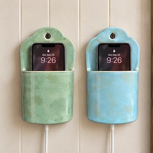 May include: Two wall-mounted ceramic phone holders, one in sage green and one in light blue, each featuring a curved top with a screw hole and a rectangular pocket holding a smartphone displaying the time 9:26 and date Sun Sep 20.