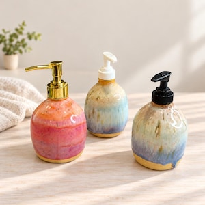 May include: Three round ceramic soap dispensers with pump mechanisms. The left dispenser is pink and orange with a gold pump. The centre dispenser is blue and tan with a white pump. The right dispenser is blue and green with a black pump.