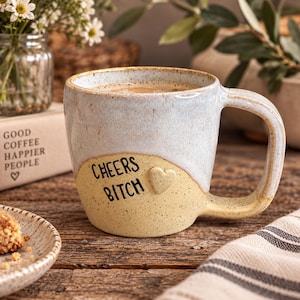 Puede incluir: Una taza de cerámica con un esmalte moteado de dos tonos, con una sección superior azul claro y una sección inferior amarilla. El texto "CHEERS BITCH" está impreso en negro en la parte frontal, junto a un pequeño detalle de cerámica en relieve.