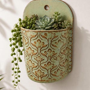 May include: A wall-mounted ceramic planter in a light green glaze with a brown embossed floral pattern. The planter features a rounded back plate with a single hole for hanging and contains a variety of green succulents, including a trailing string of pearls.