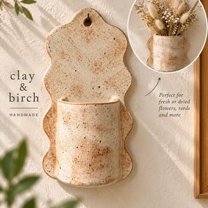 May include: A wall-mounted ceramic planter with a wavy, scalloped edge and a speckled beige finish. The piece features a central cylindrical pocket for holding items and a small hole at the top for hanging. Text on the image reads clay & birch.