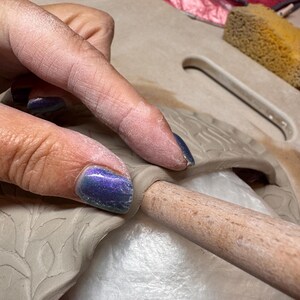 May include: Close-up of a hand working with clay. The hand is pressing a wooden tool into the clay, which is molded into a leaf design. The clay is a light beige color, and the hand has blue nail polish.