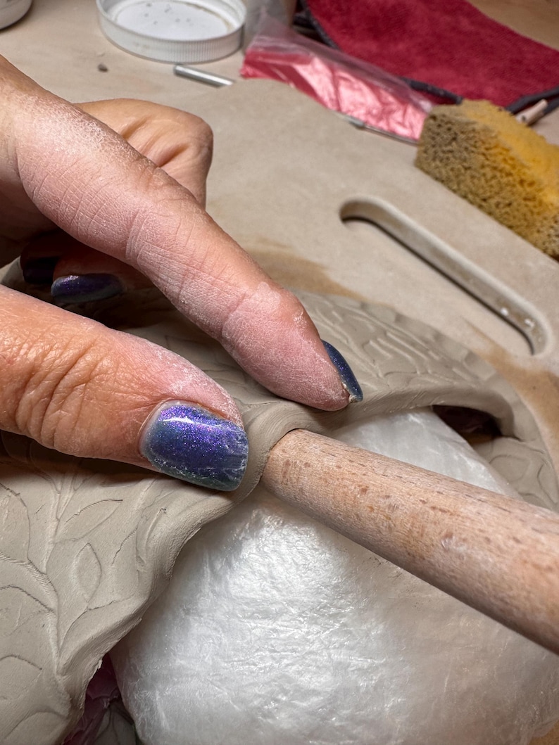 May include: Close-up of a hand working with clay. The hand is pressing a wooden tool into the clay, which is molded into a leaf design. The clay is a light beige color, and the hand has blue nail polish.