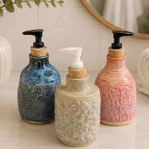 Può includere: Tre dispenser di sapone in ceramica con tappi in sughero e meccanismi a pompa. Il dispenser a sinistra è blu con un motivo topografico, quello centrale è giallo con rilievo floreale e quello a destra è rosa con un motivo a stella.