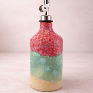 Puede incluir: Un dispensador de aceite de cer&aacute;mica con un esmalte degradado, una parte superior rosa texturizada, una secci&oacute;n central verde menta lisa con manchas circulares azul claro y una base beige s&oacute;lida. La botella tiene un vertedor de metal plateado.