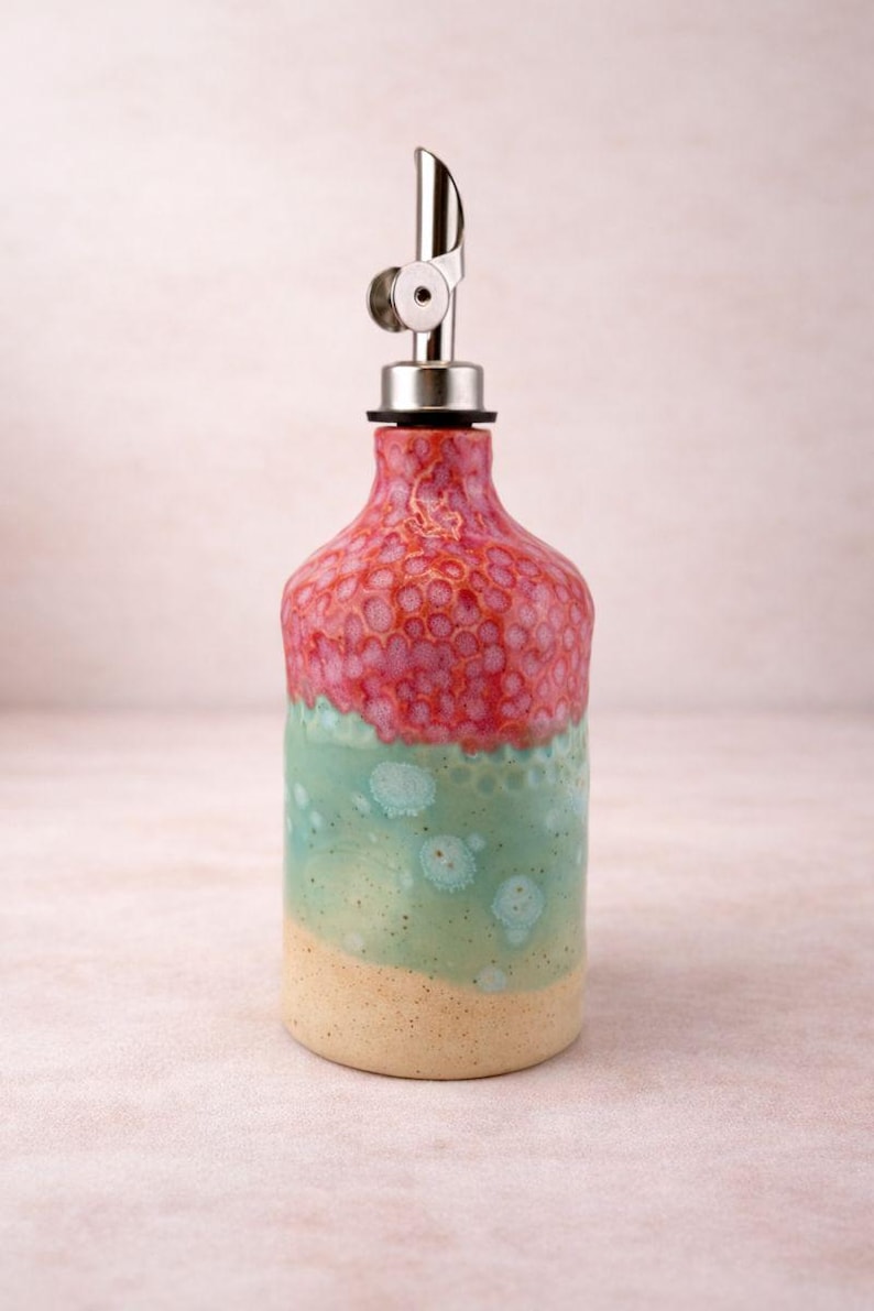 Puede incluir: Un dispensador de aceite de cer&aacute;mica con un esmalte degradado, una parte superior rosa texturizada, una secci&oacute;n central verde menta lisa con manchas circulares azul claro y una base beige s&oacute;lida. La botella tiene un vertedor de metal plateado.