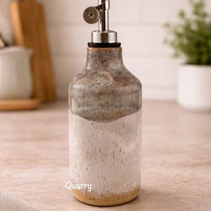 Puede incluir: Una botella dispensadora de aceite de cer&aacute;mica con un esmalte de dos tonos, con una secci&oacute;n inferior blanca moteada y una secci&oacute;n superior marr&oacute;n y gris. La botella tiene un vertedor de acero inoxidable y la palabra Quarry en blanco.