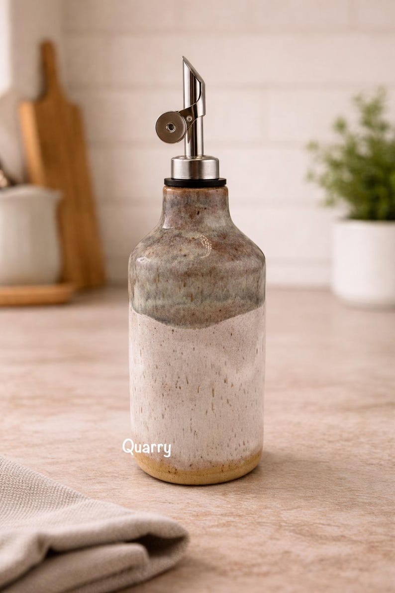 Puede incluir: Una botella dispensadora de aceite de cer&aacute;mica con un esmalte de dos tonos, con una secci&oacute;n inferior blanca moteada y una secci&oacute;n superior marr&oacute;n y gris. La botella tiene un vertedor de acero inoxidable y la palabra Quarry en blanco.