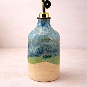 Puede incluir: Una botella dispensadora de aceite de cer&aacute;mica con un esmalte texturizado multicolor que presenta una base beige, una banda de color verde brillante y una secci&oacute;n superior de azul moteado. La botella tiene un vertedor de metal dorado con tap&oacute;n negro.