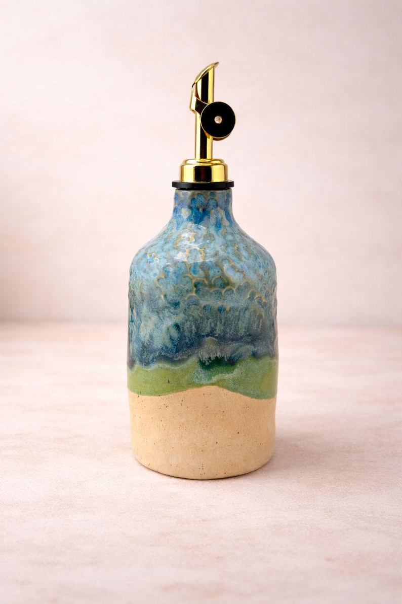 Puede incluir: Una botella dispensadora de aceite de cer&aacute;mica con un esmalte texturizado multicolor que presenta una base beige, una banda de color verde brillante y una secci&oacute;n superior de azul moteado. La botella tiene un vertedor de metal dorado con tap&oacute;n negro.