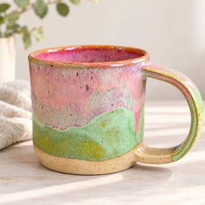 Puede incluir: Una taza de cerámica con una base beige moteada que transiciona hacia una sección superior esmaltada en tonos verde y rosa vibrantes. La taza presenta un asa ancha y resistente, junto con un borde redondeado y suave de aspecto artesanal.