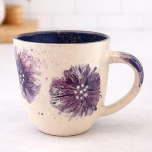 Peut inclure: Mug en céramique crème avec deux grandes fleurs violettes. L'intérieur est bleu foncé avec des mouchetures dorées. La poignée et les détails floraux sont violets. Un objet de boisson unique.