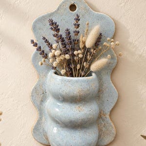 May include: A light blue ceramic wall pocket with a wavy, scalloped edge and a speckled finish. The piece features a hole at the top for hanging and contains a dried floral arrangement of purple lavender, white baby's breath, and tan bunny tails.