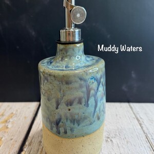 Puede incluir: Un dispensador de aceite de cer&aacute;mica con un vertedor met&aacute;lico. El cuerpo presenta una base beige moteada y un acabado de esmalte de goteo azul y verde. El texto Muddy Waters est&aacute; impreso a la derecha del cuello de la botella.
