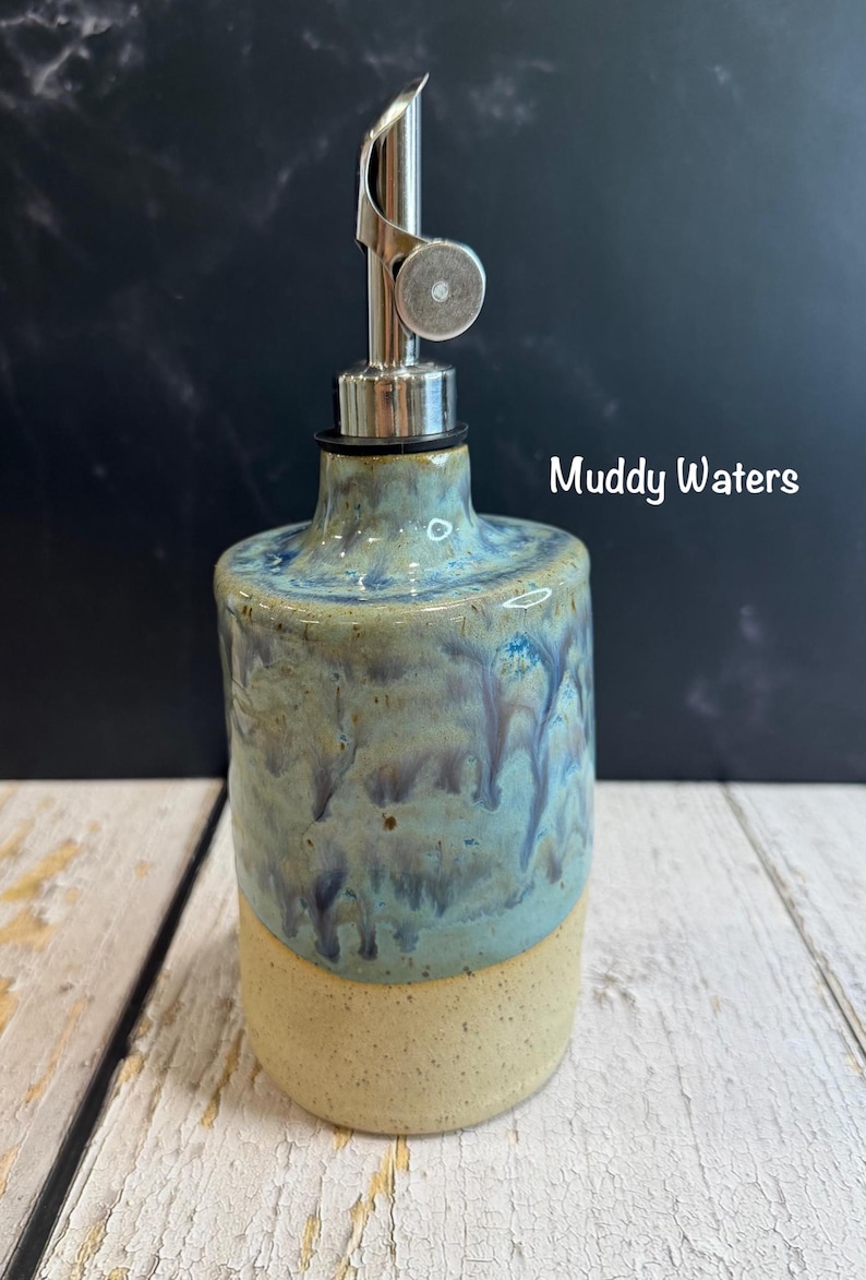 Puede incluir: Un dispensador de aceite de cer&aacute;mica con un vertedor met&aacute;lico. El cuerpo presenta una base beige moteada y un acabado de esmalte de goteo azul y verde. El texto Muddy Waters est&aacute; impreso a la derecha del cuello de la botella.