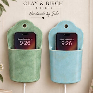 May include: Two wall-mounted ceramic phone holders in sage green and light blue, each featuring a rounded top with a circular hole for hanging. A smartphone is placed inside each holder, with a white charging cable extending from the bottom.