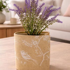Peut inclure: Pot en céramique beige rectangulaire avec un motif d'oiseau et de fleurs blanches, contenant une plante de lavande aux fleurs violettes et aux tiges vertes. Posé sur une table en bois.