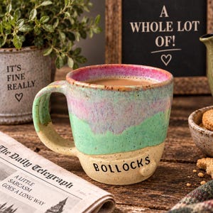 Peut inclure: Une tasse en céramique avec une finition mouchetée présentant un dégradé de couleurs rose, vert et beige. Le mot BOLLOCKS est imprimé en lettres capitales noires sur la section beige inférieure. La tasse possède une anse courbée et un large bord.
