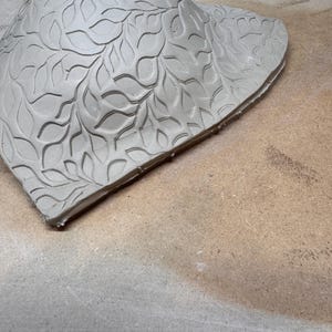 May include: A light gray, unfinished ceramic piece with a leaf pattern carved into the surface. The clay is in a curved, irregular shape, possibly a decorative bowl or wall hanging. The edges are rough, showing the handmade nature of the item.