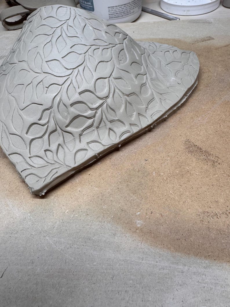 May include: A light gray, unfinished ceramic piece with a leaf pattern carved into the surface. The clay is in a curved, irregular shape, possibly a decorative bowl or wall hanging. The edges are rough, showing the handmade nature of the item.