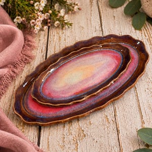 May include: Two oval ceramic trays with scalloped edges, featuring a glossy glaze that transitions from a deep brown rim to a vibrant pink and soft yellow center. The trays are nested together, showcasing a smooth, reflective surface finish.